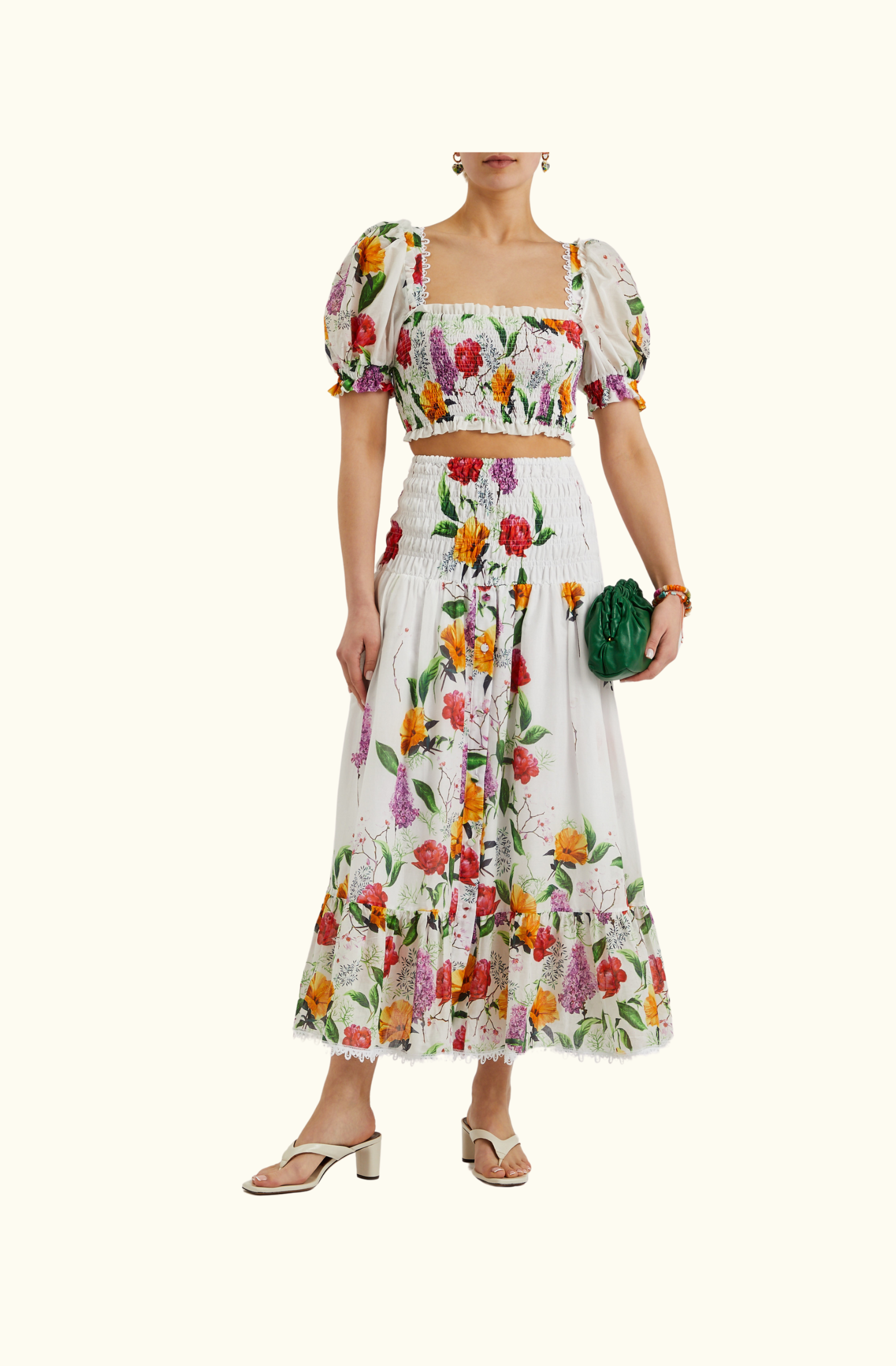 Model wearing the Charo Ruiz Ibiza co-ord set with a square-neck floral crop top and a smocked high-waist floral maxi skirt, styled with white heeled sandals and a green clutch bag.