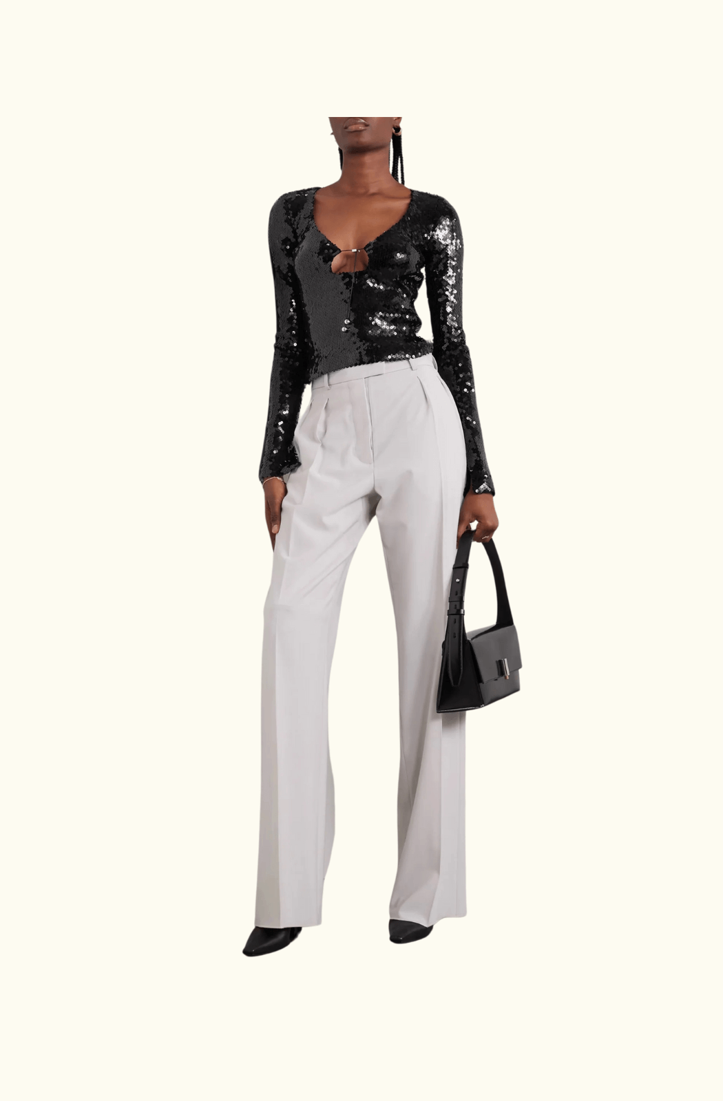 A model wearing the black sequinned stretch-tulle top by 16Arlington styled with high-waisted white tailored trousers, black pointed-toe heels, and a black structured handbag, posing against a minimal studio setting with concrete accents.