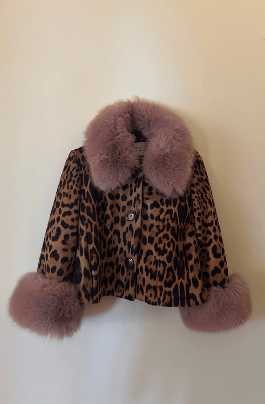 Charlotte Simone Cropped Edie Jacket in leopard print hanging against a plain wall. The jacket features a plush faux fur collar and matching faux fur cuffs in a soft pink shade. It has a cropped silhouette, a structured fit, and a button-up front closure. The detachable fur trims allow for customisation with pink, black, or no fur at all.