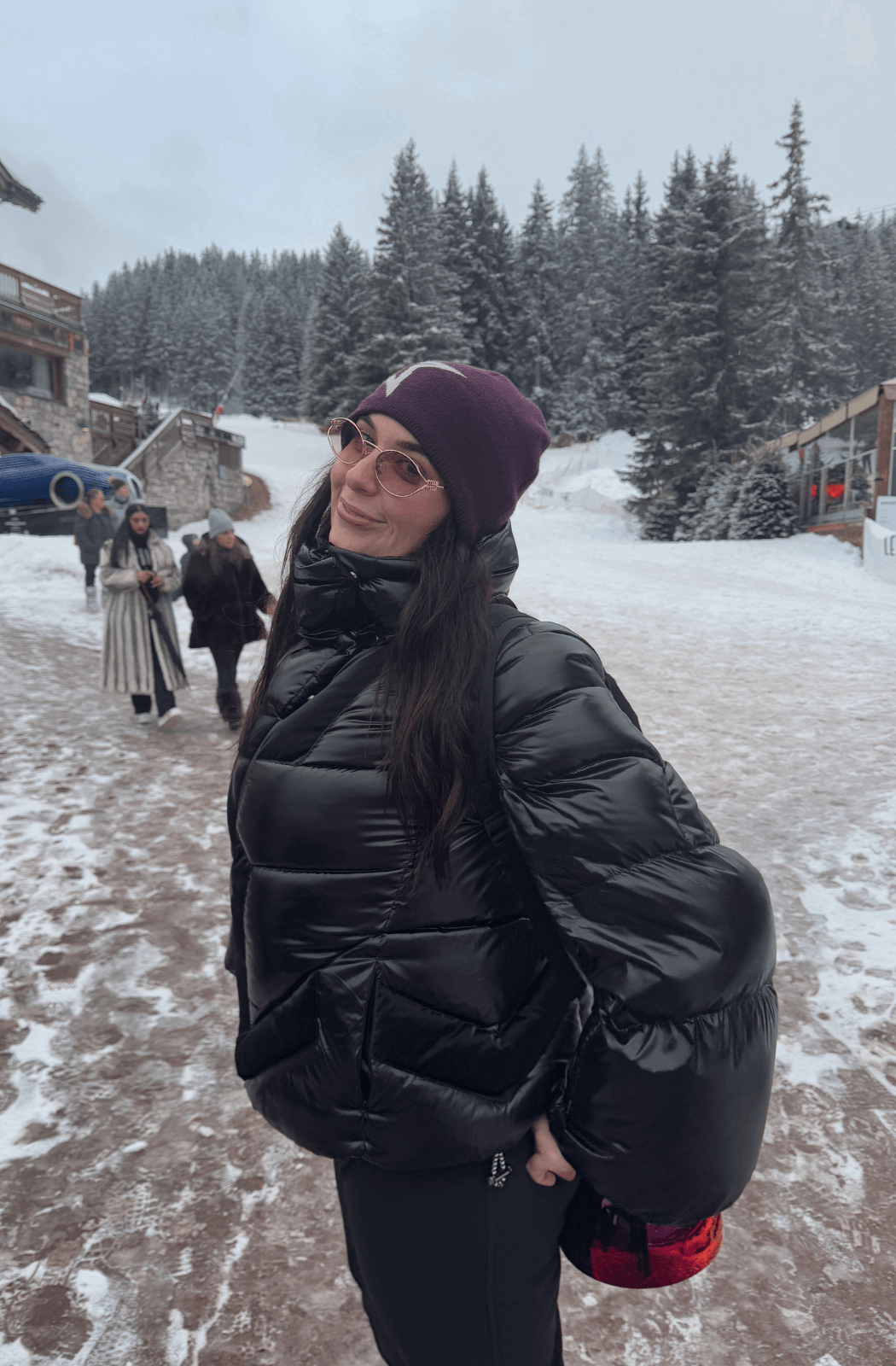 A mid-length shot of the Ariel Down Bomber Jacket in black, worn in a snowy alpine setting. The glossy, quilted fabric reflects light, emphasizing its voluminous silhouette. The oversized fit, paired with a high-neck collar and puffed sleeves, offers both warmth and an effortlessly chic aesthetic.