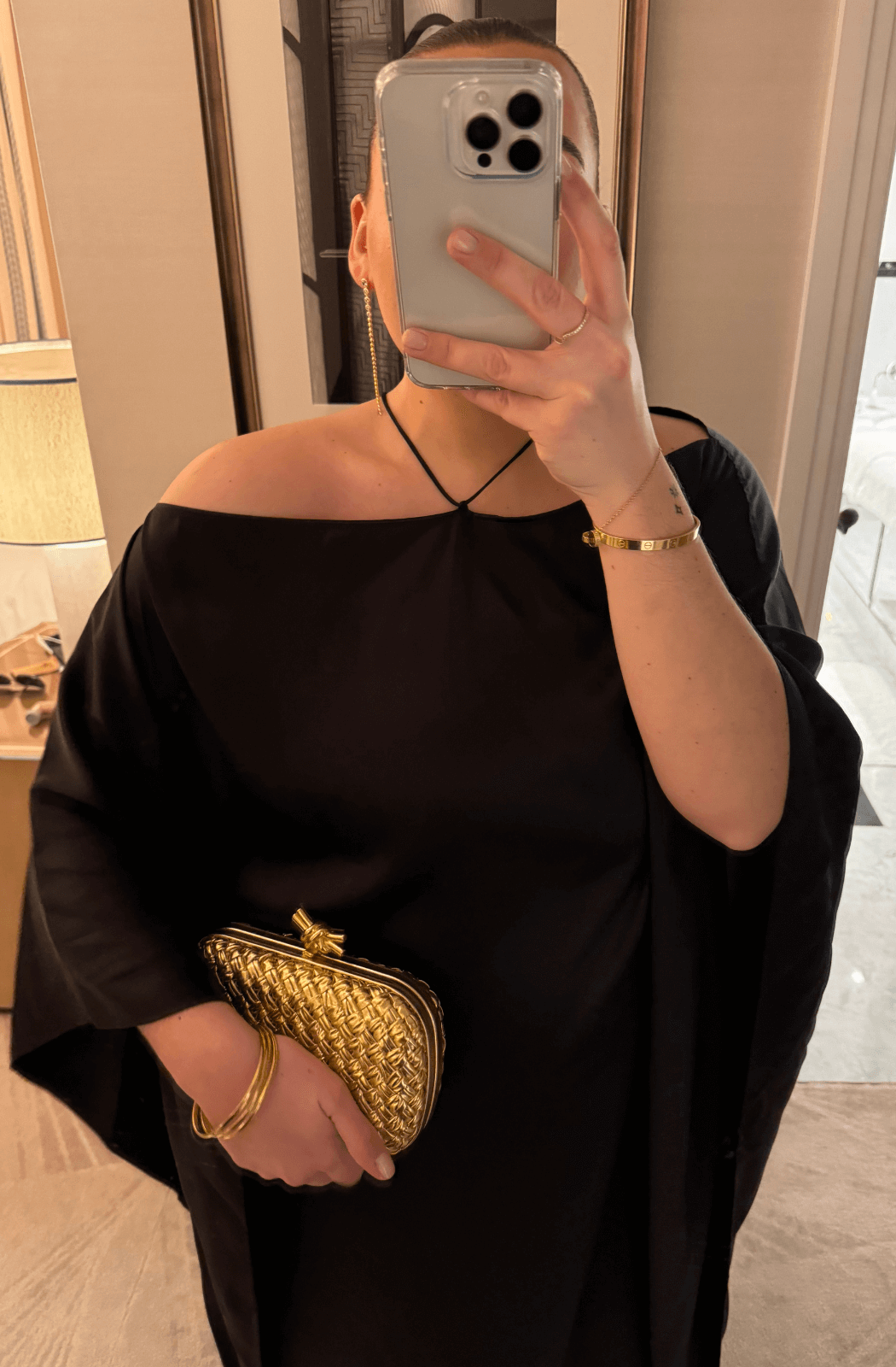 A close-up photo of a person wearing the Taller Marmo Sza silk maxi dress, showing off the halter neckline and off-the-shoulder draping. The individual holds a gold woven clutch and accessorizes with gold jewelry, adding a touch of elegance to the look.